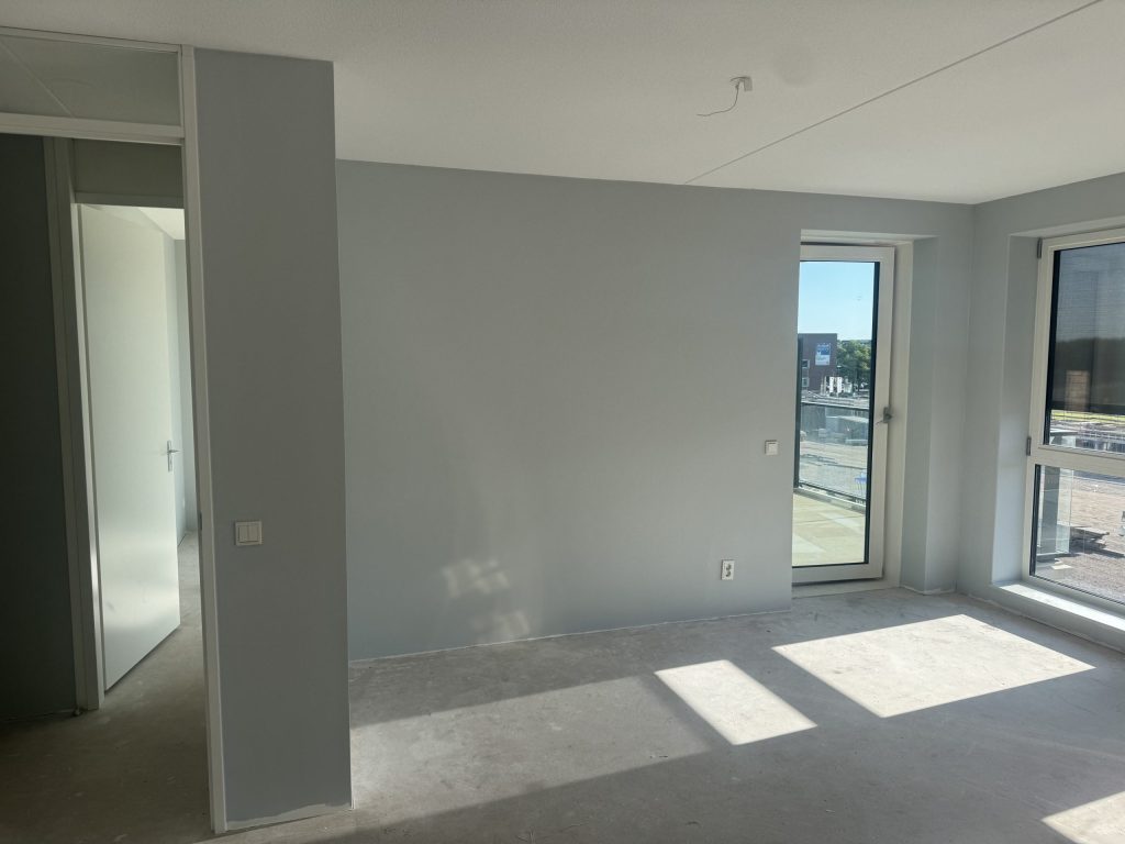 Kale woonkamer in een nieuw appartement met lichtgrijze wanden, een balkondeur en een betonvloer, nog zonder behang