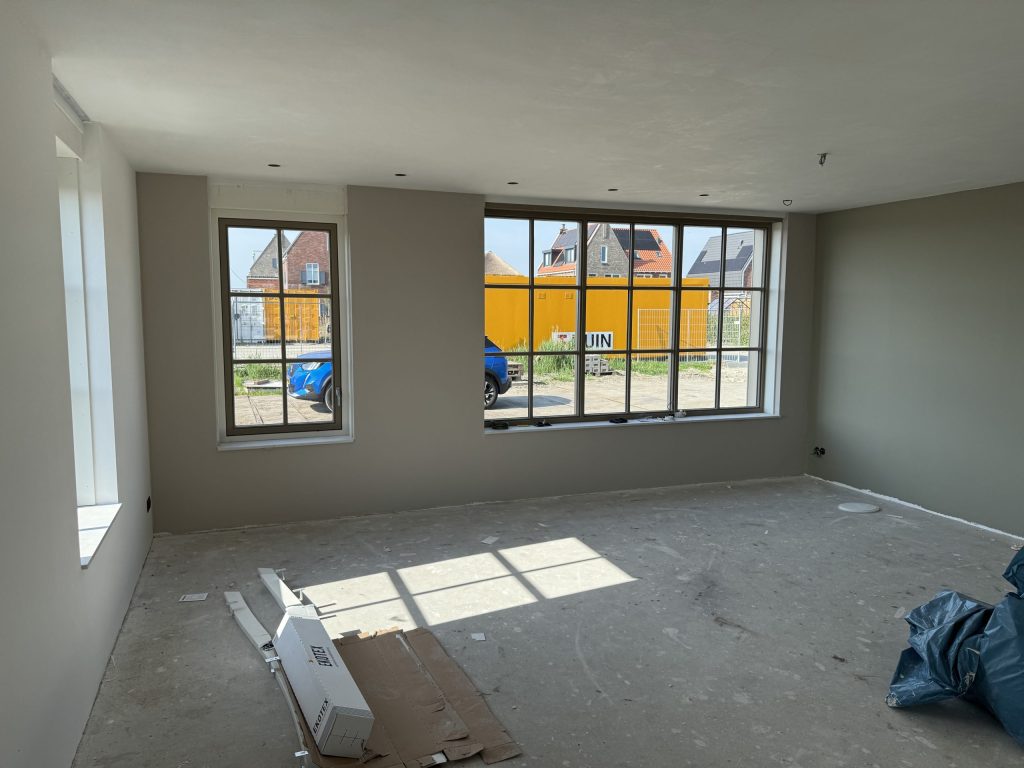 Nieuw opgeleverde woonkamer met taupe gesausde wanden, grote ramen met roedes en een kale betonvloer