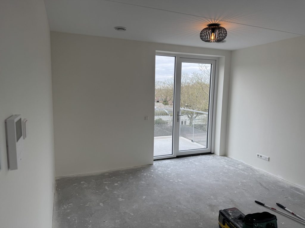 Woonkamer van een nieuw appartement met witte wanden, een balkondeur met glazen hek en een betonvloer