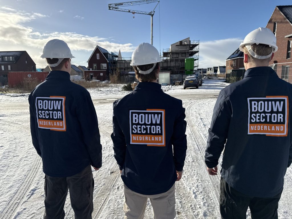 Drie medewerkers van Bouwsector Nederland met witte helmen op de rug gezien, op een besneeuwde bouwstraat met nieuwe woningen en een kraan